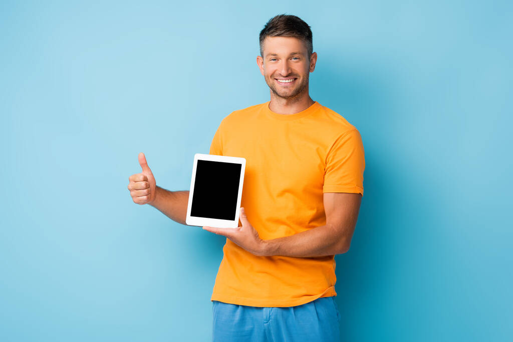 Tişörtlü mutlu adam elinde boş ekranlı dijital tablet tutuyor ve mavi ekranda başparmağını gösteriyor. - Fotoğraf, Görsel