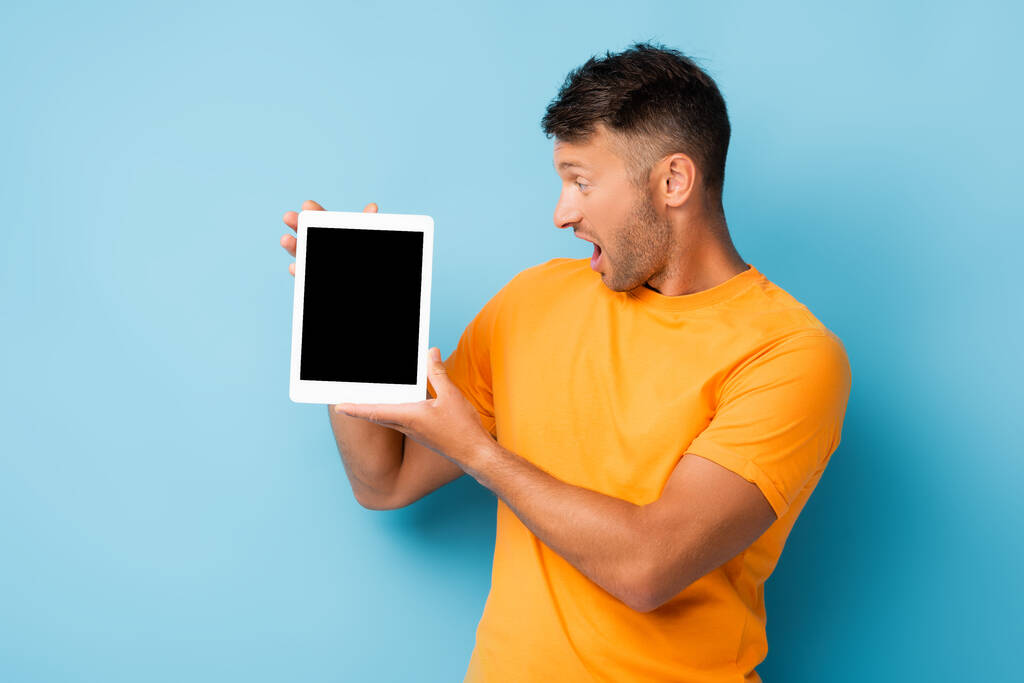 Überraschter Mann im T-Shirt blickt auf digitales Tablet mit leerem Bildschirm auf blauem Hintergrund - Foto, Bild