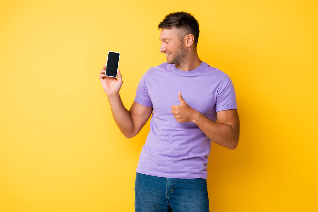 glücklicher Mann hält Smartphone mit leerem Bildschirm und zeigt Daumen hoch auf gelb  - Foto, Bild