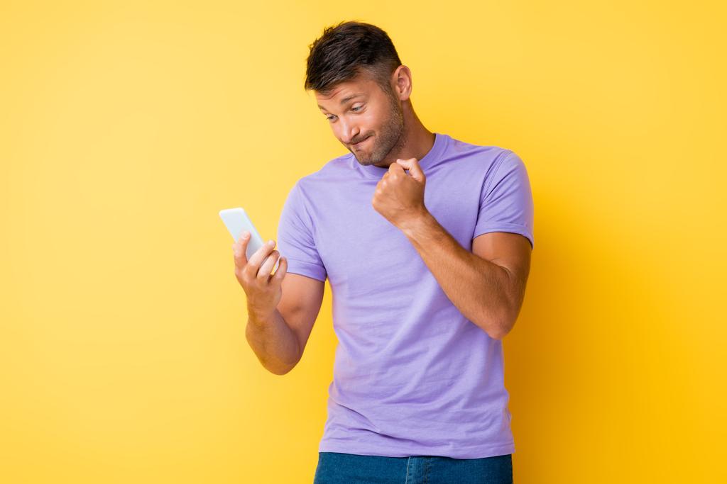 недовольный мужчина показывает сжатый кулак и смотрит на смартфон на желтом - Фото, изображение
