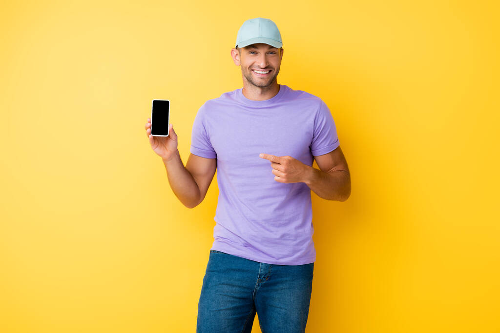 happy man in blue cap pointing with finger at smartphone with blank screen on yellow - Photo, Image