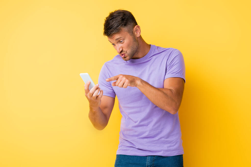 displeased man pouting lips while pointing with finger at smartphone on yellow - Photo, Image