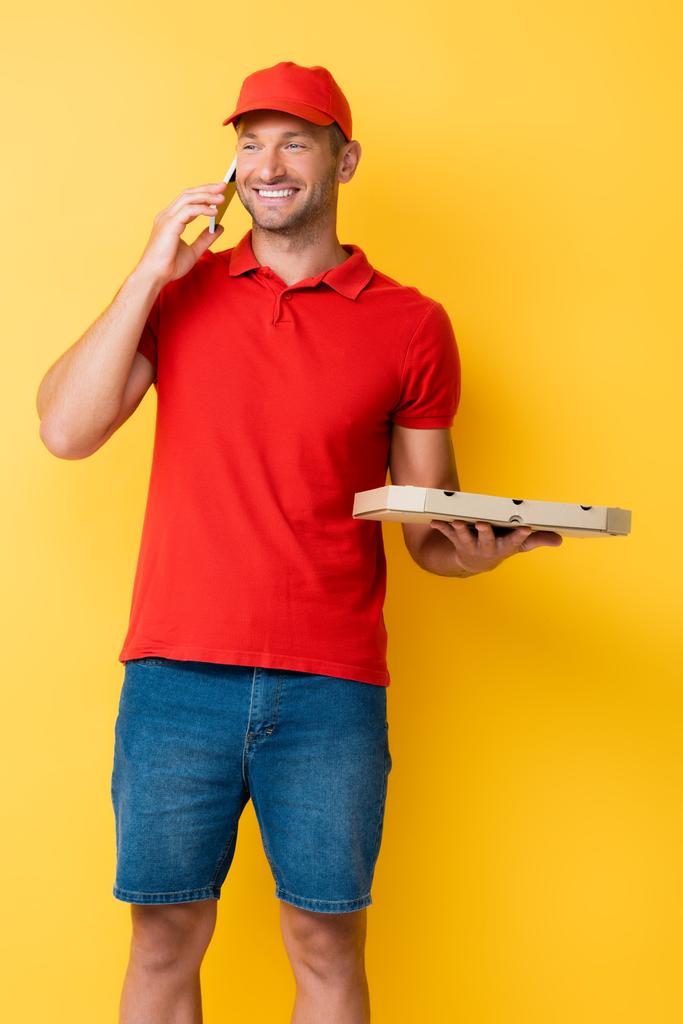 hombre de entrega feliz en caja de sujeción de tapa roja con sabrosa pizza y hablando en el teléfono inteligente en amarillo - Foto, imagen