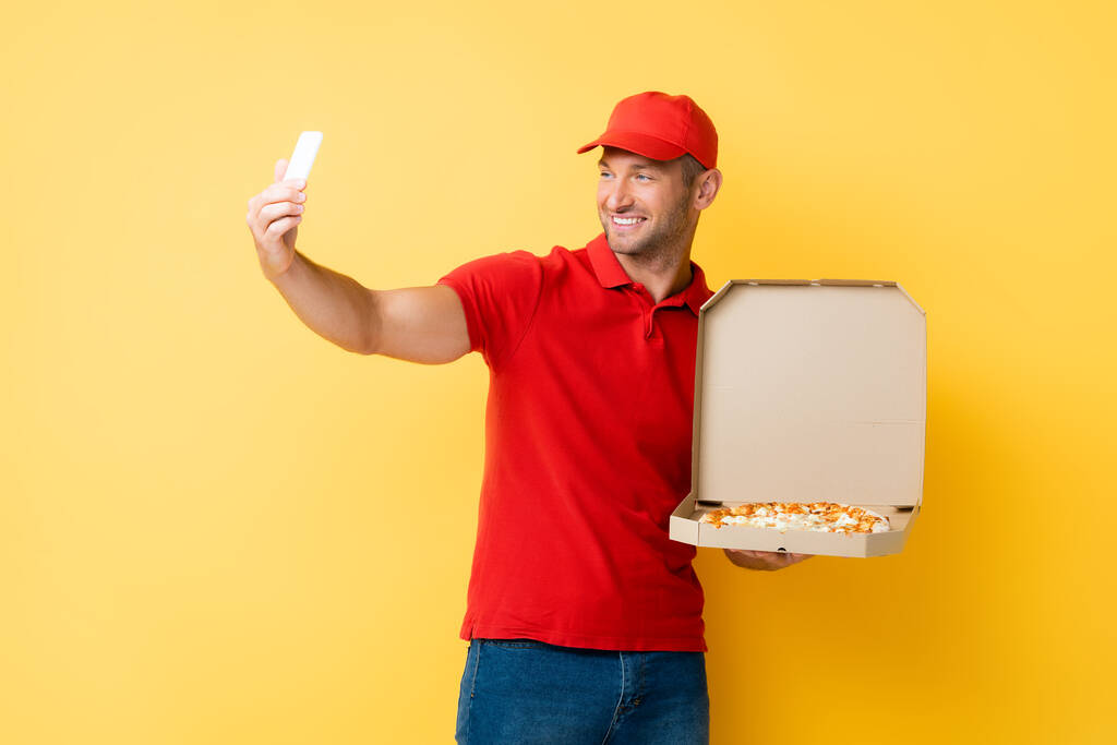 Lächelnder Lieferant in roter Mütze mit leckerer Pizza und Selfie auf gelb - Foto, Bild