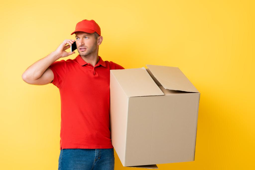 Kırmızı şapkalı teslimatçı elinde karton kutuyla sarıda akıllı telefondan konuşuyor. - Fotoğraf, Görsel