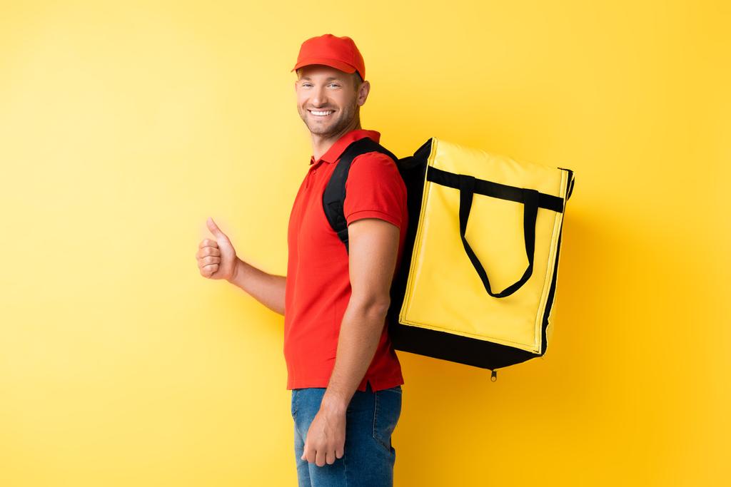 alegre repartidor hombre llevando mochila con orden y mostrando el pulgar hacia arriba en amarillo - Foto, Imagen