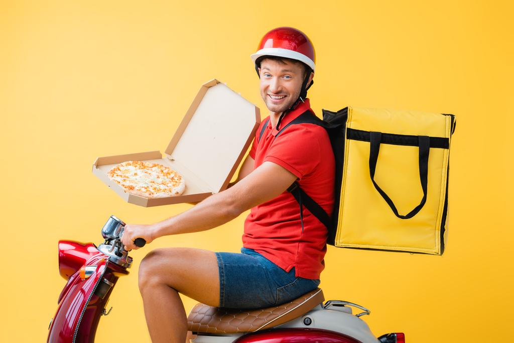happy delivery man with backpack riding red scooter and holding pizza in carton box on yellow - Photo, Image