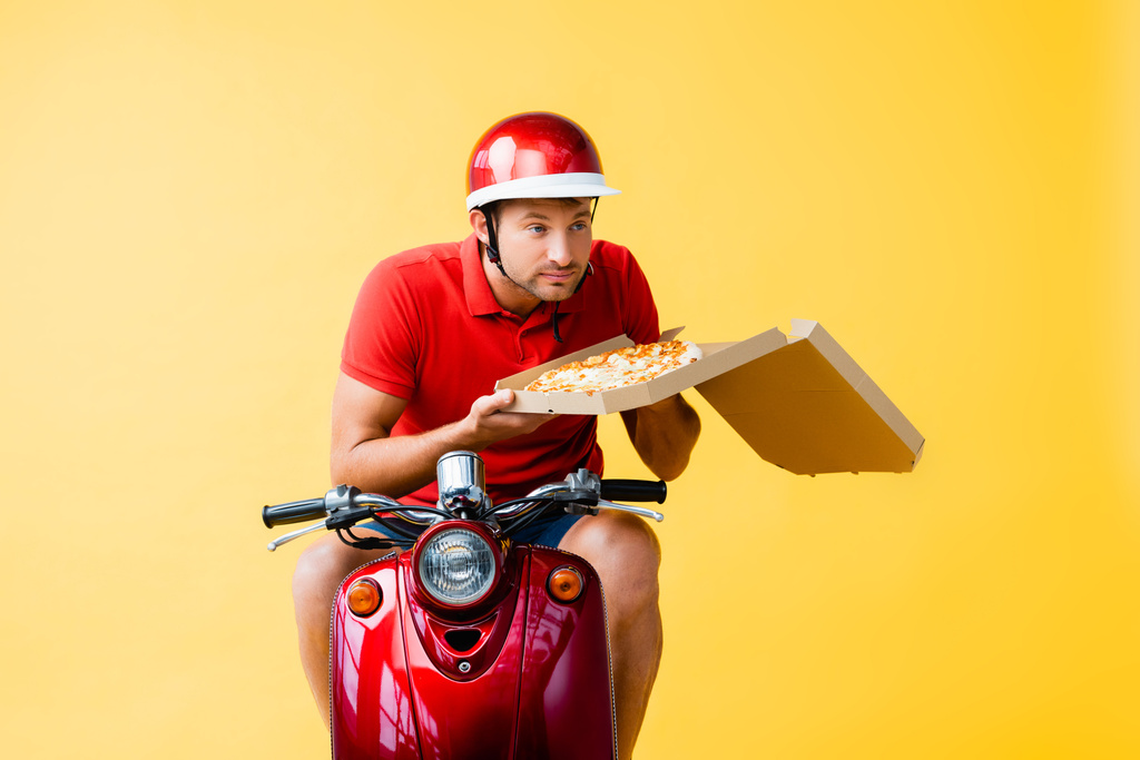 repartidor en casco montando scooter rojo y oliendo pizza en caja de cartón en amarillo - Foto, Imagen