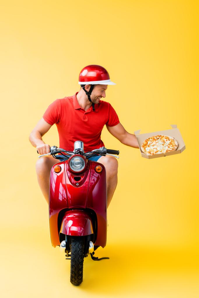 livreur souriant dans un casque équitation scooter rouge et tenant la pizza dans une boîte en carton sur jaune - Photo, image