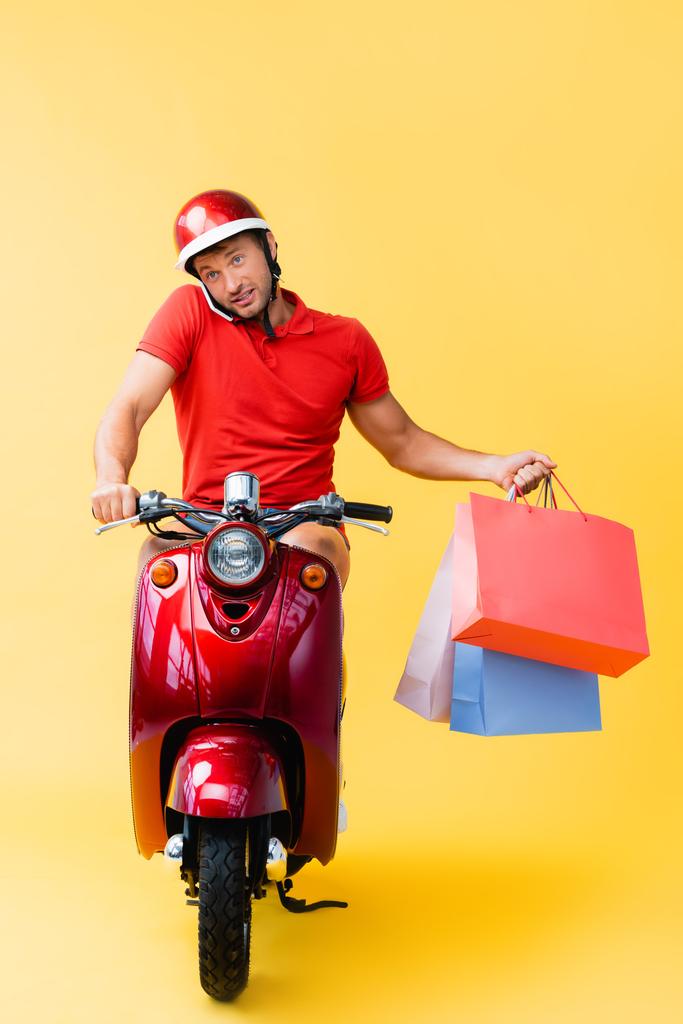 repartidor en casco a caballo scooter y teléfono inteligente hablando mientras sostiene bolsas de compras en amarillo  - Foto, imagen