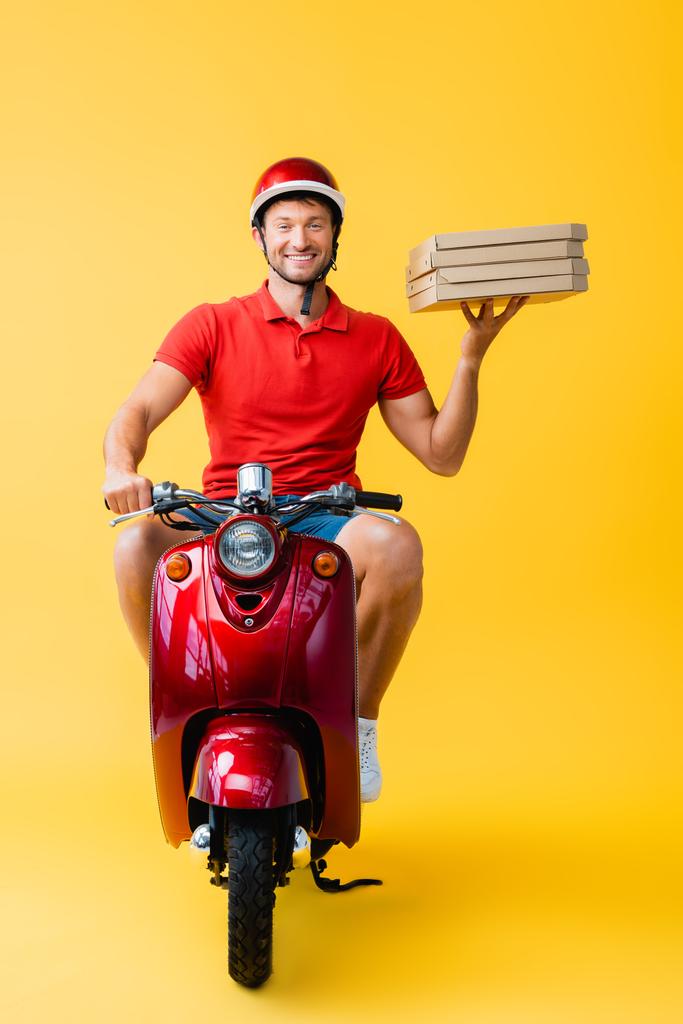幸せな配達人でヘルメットに乗るスクーターと保持カートンピザボックスで黄色  - 写真・画像