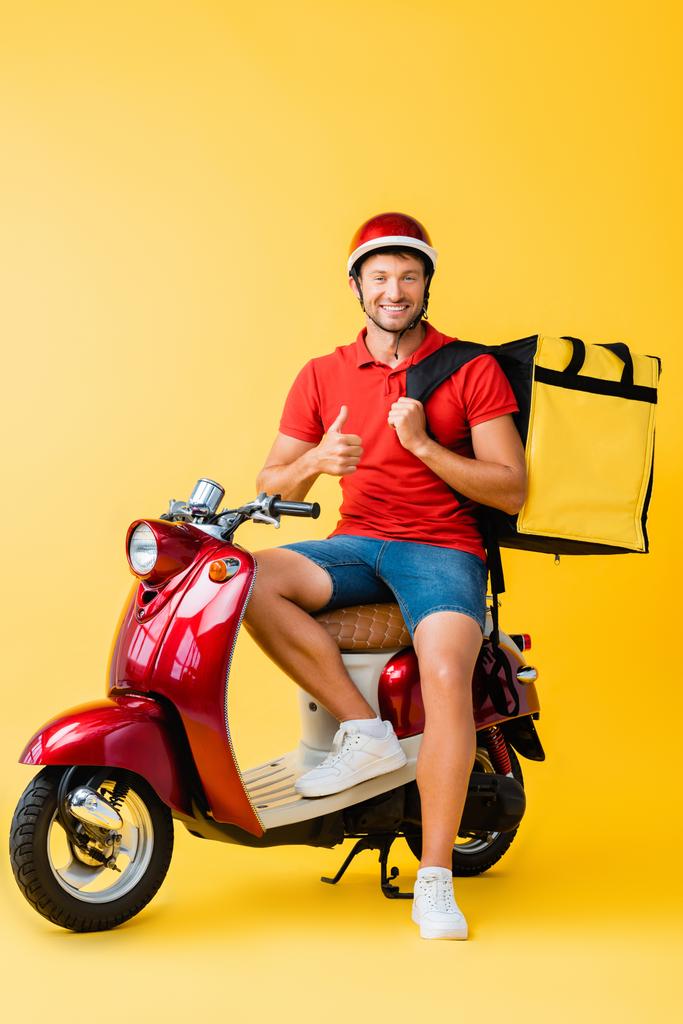 счастливый доставщик с рюкзаком сидит на красном скутере и показывает большой палец вверх на желтом - Фото, изображение