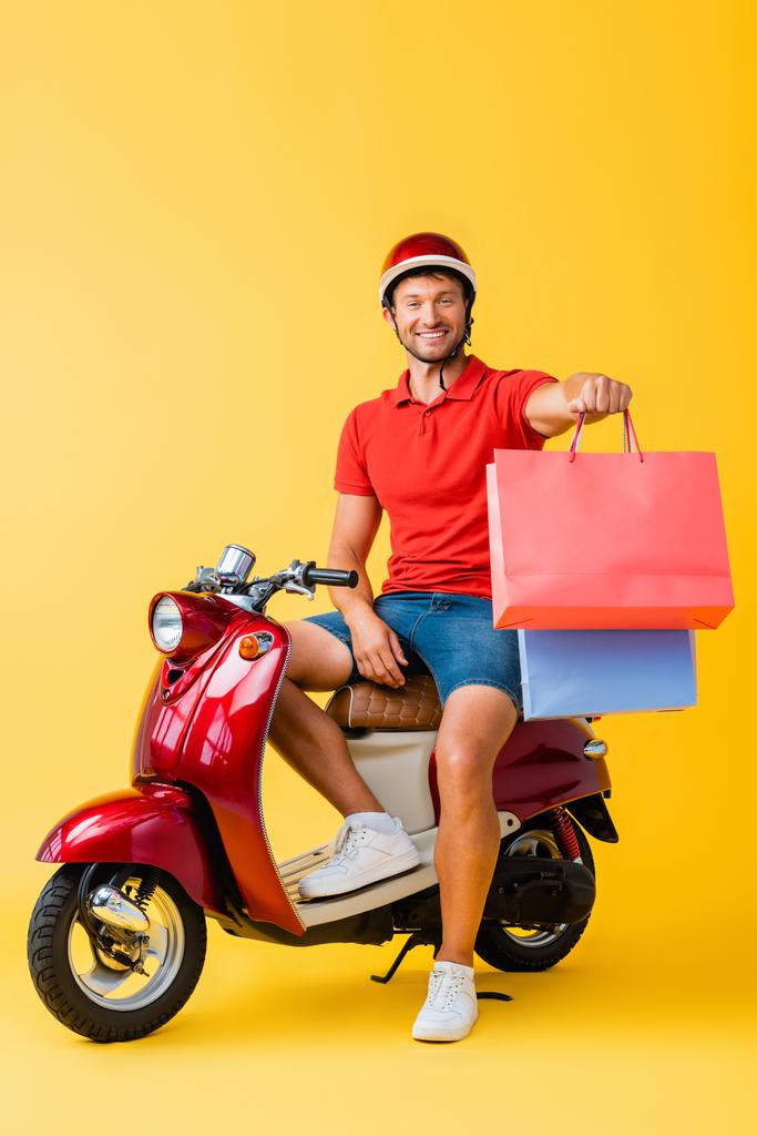 счастливый доставщик в шлеме, сидящий на скутере и держащий сумки с покупками на желтом  - Фото, изображение
