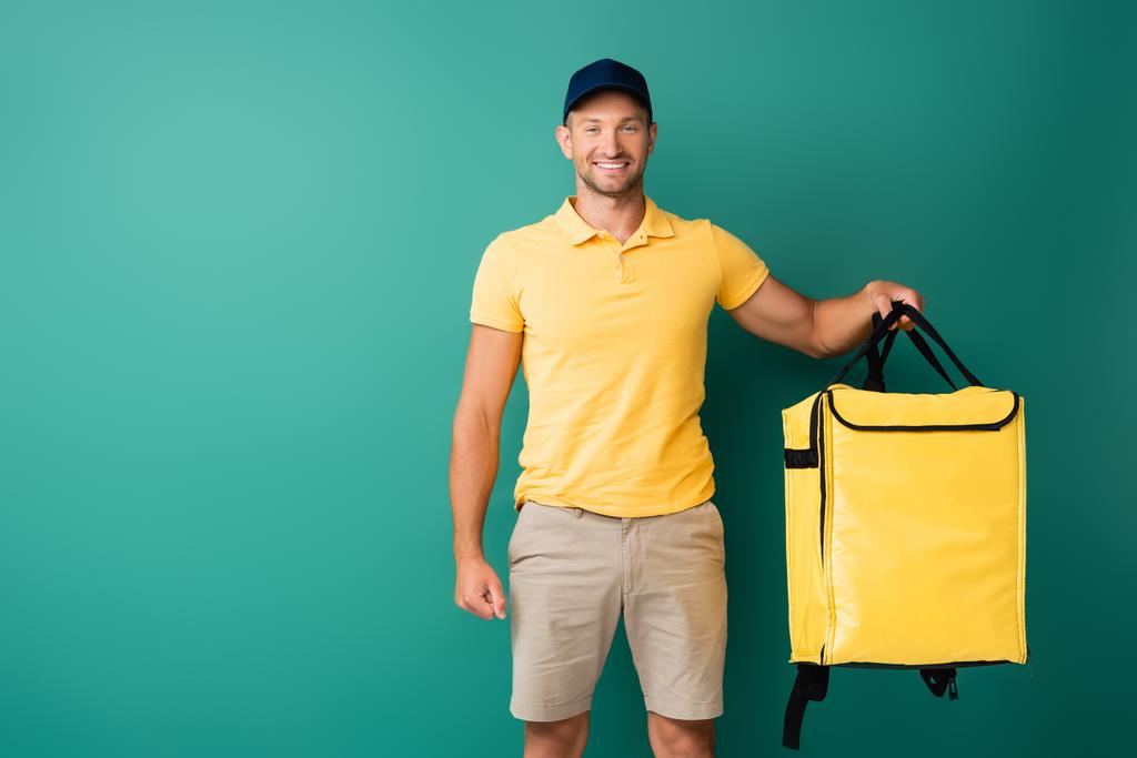 homem de entrega alegre carregando mochila amarela e sorrindo no azul - Foto, Imagem