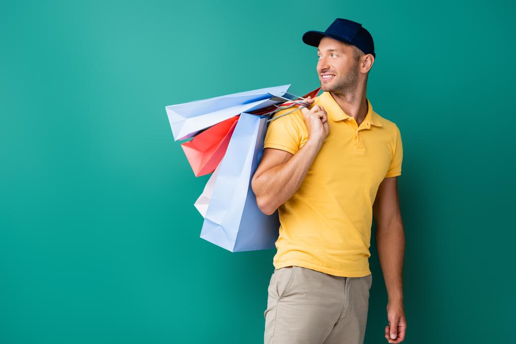 Şapkalı neşeli teslimatçı alışveriş torbaları taşıyor ve maviye bakıyor. - Fotoğraf, Görsel