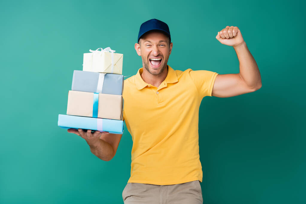 podekscytowany dostawca w czapce trzymając pakowane prezenty na niebiesko - Zdjęcie, obraz