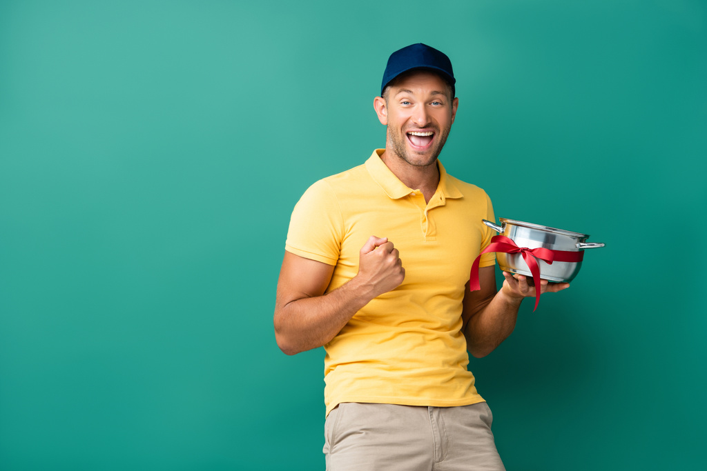 smiling delivery man in cap holding saucepan with ribbon on blue  - Photo, Image