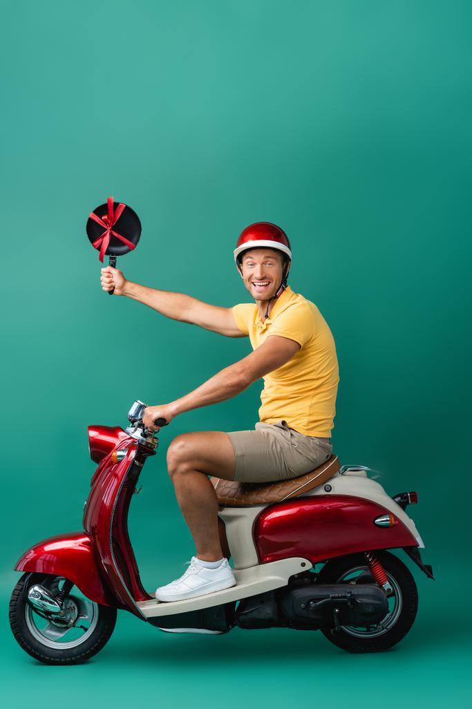 щасливий чоловік доставки в шолом їде на мотоциклі і тримає сковороду зі стрічкою на синьому
 - Фото, зображення