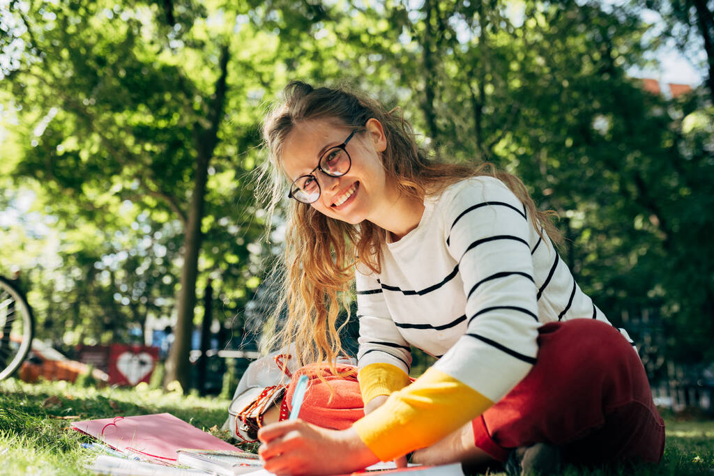 Студентка в повседневной одежде, в очках, сидит на зеленой траве в университетском городке и учится на улице. Улыбающаяся молодая женщина учится в парке. - Фото, изображение