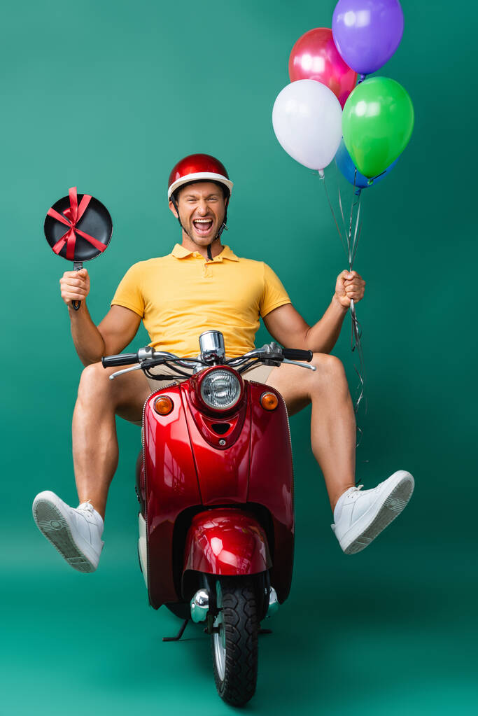 uomo consegna eccitato in casco cavalcando scooter tenendo palloncini e nuova padella su blu - Foto, immagini