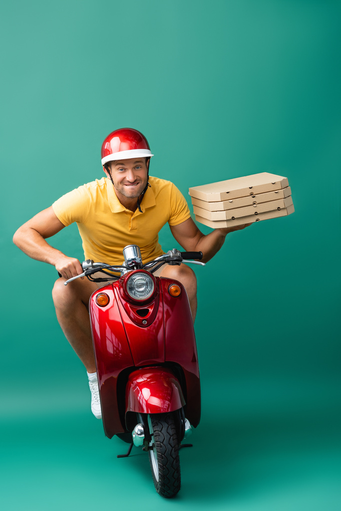 Zusteller im Helm beißt beim Rollerfahren auf die Lippen, während er Pizzakartons auf blau hält - Foto, Bild