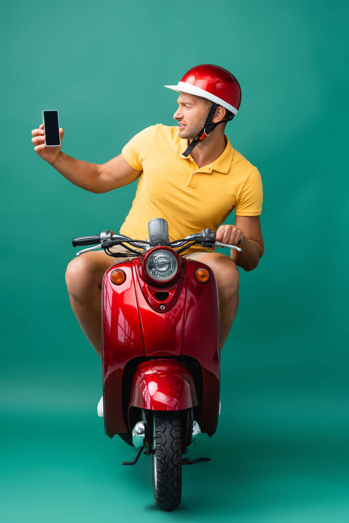 青い画面で空白の画面でスマートフォンを保持しながらヘルメットに乗るスクーターの配達人 - 写真・画像