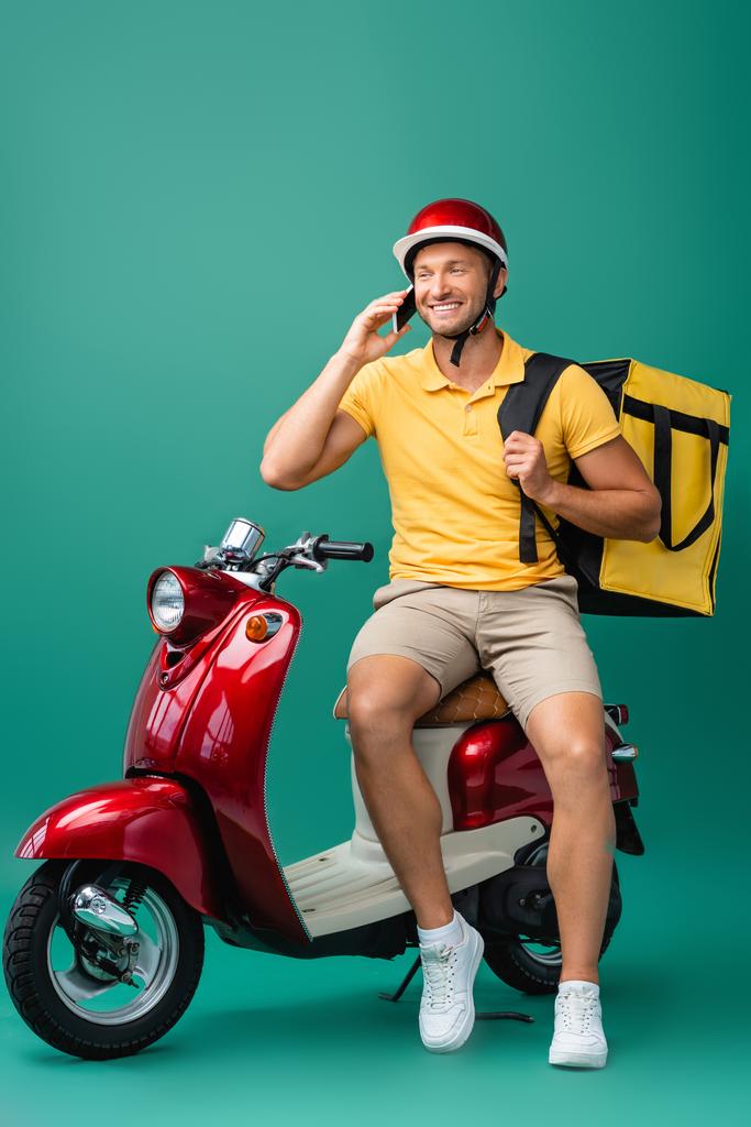 ブルーのスクーターの近くのスマートフォンで話すバックパックを持った陽気な配達の男 - 写真・画像