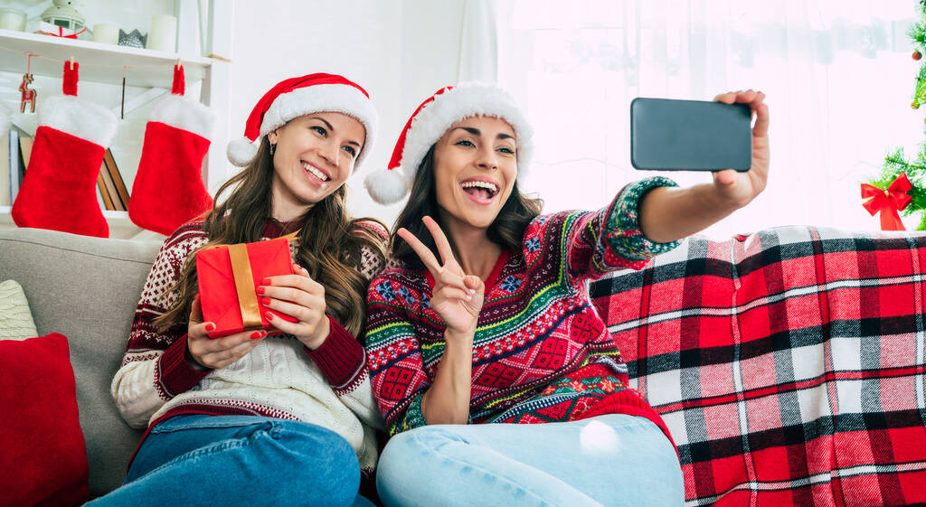 Gelukkig Nieuwjaar en Vrolijk Kerstfeest. Twee mooie opgewonden jonge zussen of vrienden in Santa hoeden maken selfie foto op de smartphone op de bank met kerst geschenkdozen. - Foto, afbeelding