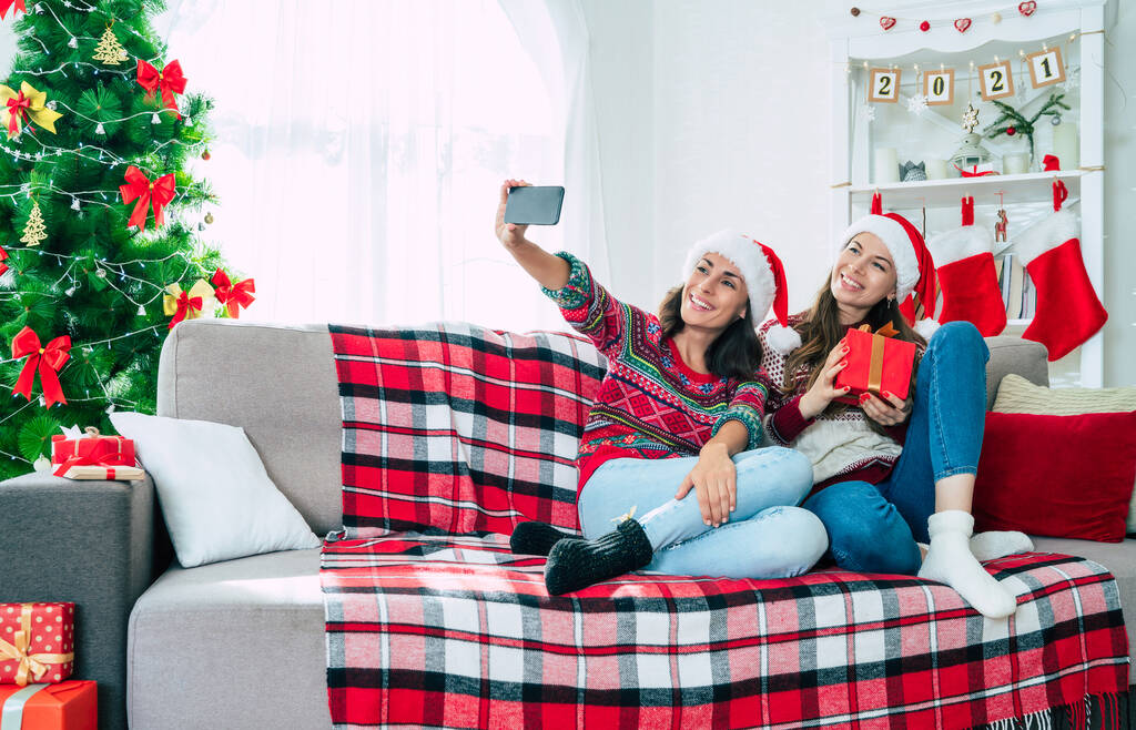 Καλή Χρονιά και Καλά Χριστούγεννα. Δύο όμορφες ενθουσιασμένες νεαρές αδελφές ή φίλοι με τα καπέλα Σάντα κάνουν selfie φωτογραφία στο smartphone στον καναπέ με κουτιά δώρων Χριστουγέννων. - Φωτογραφία, εικόνα