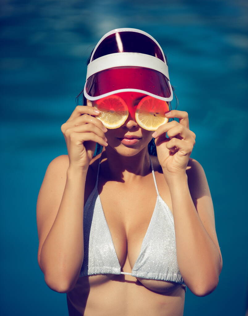 Mooie vrouw in een zilveren badpak en een rode zonneklep met citroenschijfjes staat in het water in het zwembad - Foto, afbeelding