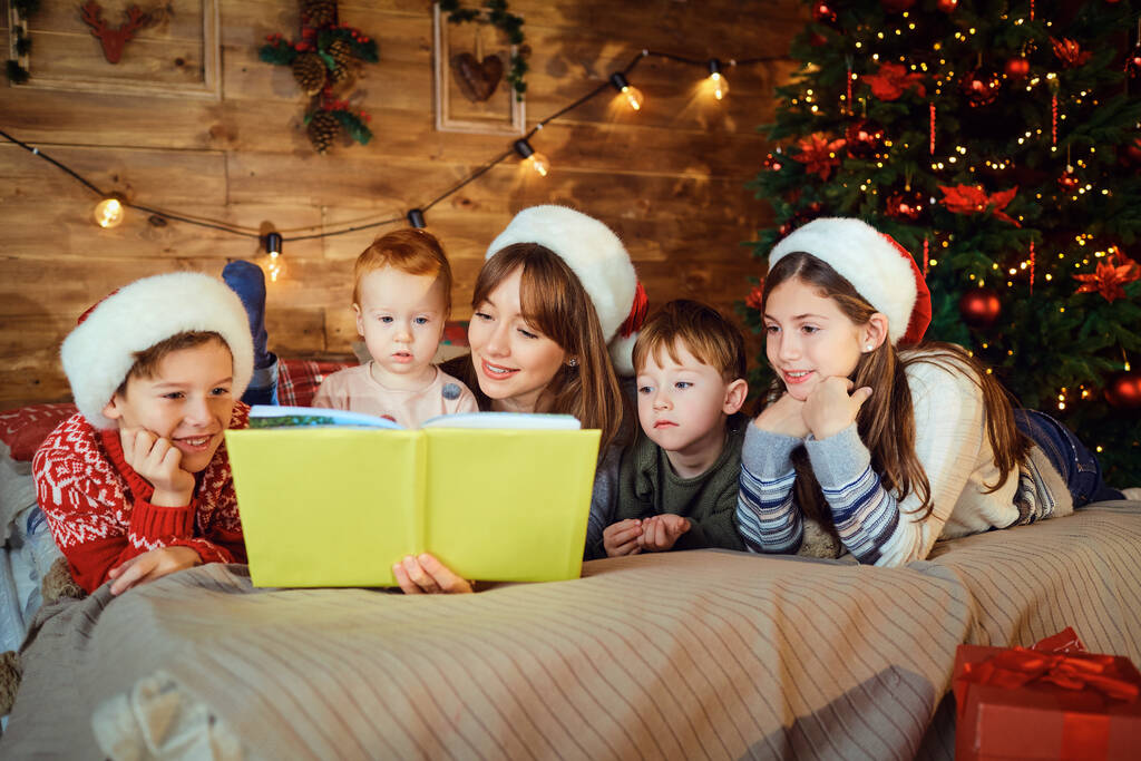 La mamma legge un libro con i bambini sdraiati sul letto in camera con albero a Natale. - Foto, immagini