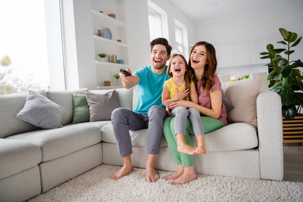 Täysikokoinen kuva positiivisia kolme ihmistä äiti isä esiteini pieni lapsi tyttö istua mukavasti sohvalla levätä katsella komedia sarjakuva kaukosäädin kytkin ilo nauraa talossa sisätiloissa - Valokuva, kuva