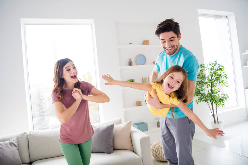 Φωτογραφία από θετικά χαρούμενα τρία άτομα παίζουν το παιχνίδι μπαμπάς μεταφέρουν μικρό παιδί που κρατά τα χέρια πετούν αεροπλάνο μητέρα αισθάνονται χαρά συναισθήματα στο σπίτι σε εσωτερικούς χώρους - Φωτογραφία, εικόνα