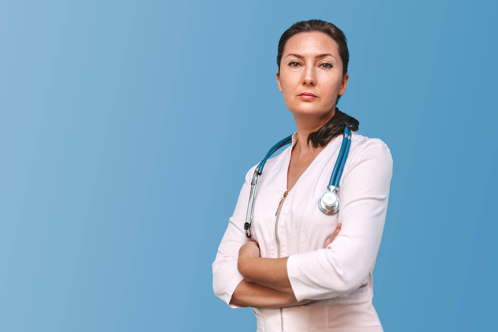Portrait d'une femme médecin adulte sérieuse aux bras croisés, regardant la caméra. Isolé sur fond bleu - Photo, image