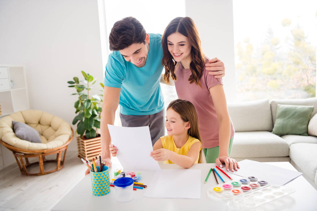 Photo of positive cheerful three people small kid girl sit table show mommy daddy beautiful painted picture paper in house indoors - Photo, Image