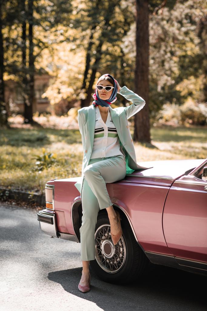 Mulher elegante em óculos de sol de pé perto do carro vintage na estrada  - Foto, Imagem
