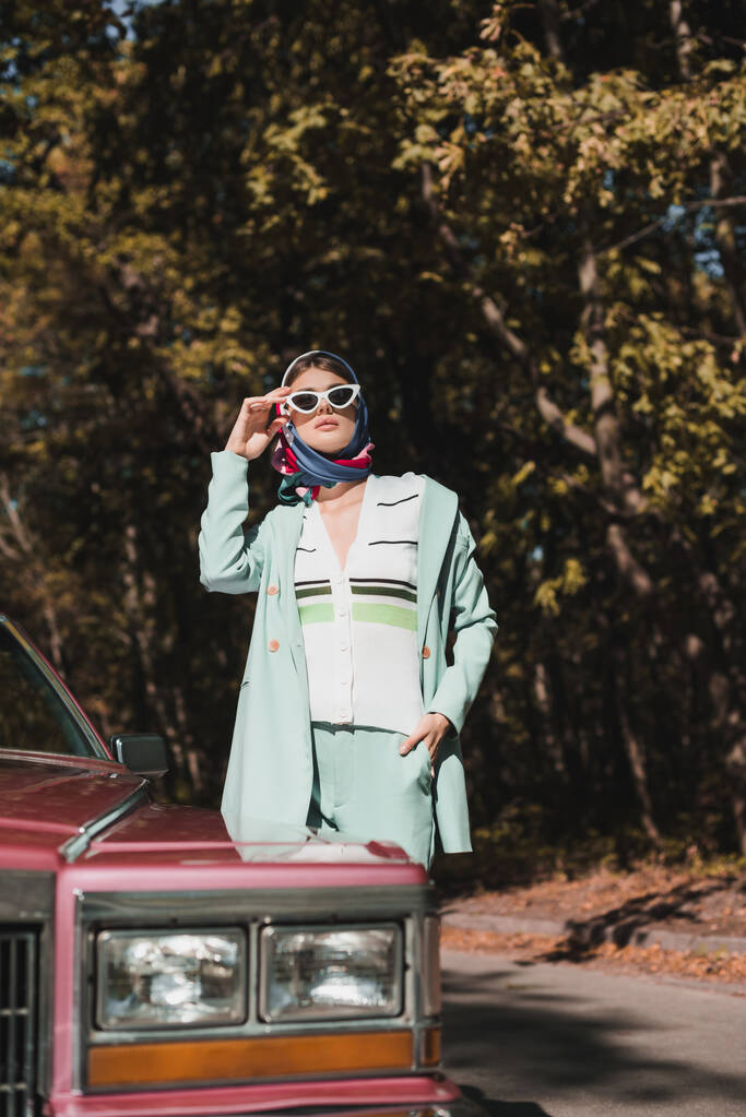 Stylowa kobieta w chustce dotykająca okularów przeciwsłonecznych w pobliżu samochodu na niewyraźnym pierwszym planie na drodze  - Zdjęcie, obraz