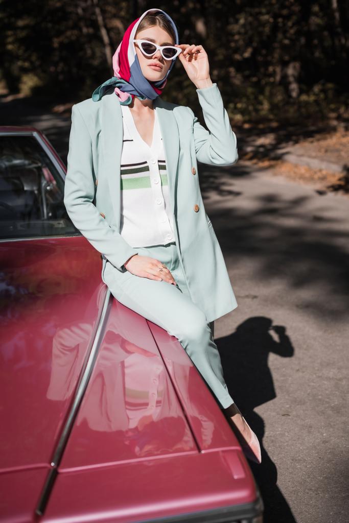 Mujer joven en gafas de sol sentada en coche vintage con la carretera en el fondo  - Foto, imagen