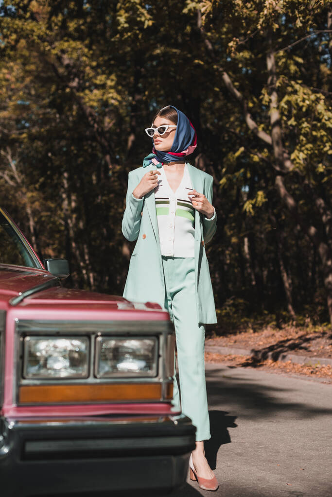 Mujer de moda de pie cerca de auto retro en primer plano borroso en la carretera  - Foto, imagen