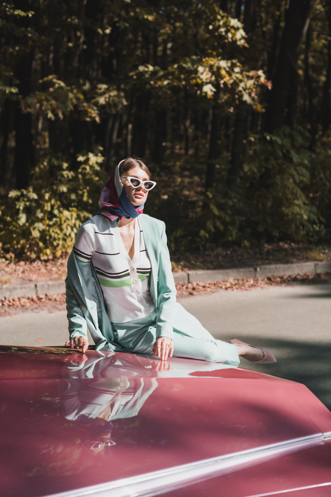 Κομψή γυναίκα με γυαλιά ηλίου κάθεται στον προφυλακτήρα του αυτοκινήτου αντίκα σε εξωτερικούς χώρους  - Φωτογραφία, εικόνα