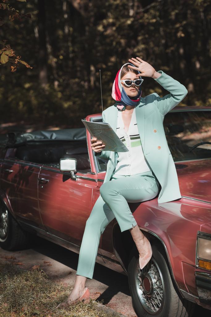 Стильная женщина в солнцезащитных очках держит карту и смотрит в сторону от винтажного автомобиля на дороге  - Фото, изображение