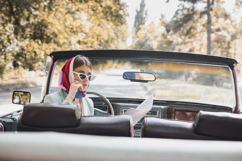 Mujer pensativa en gafas de sol mirando el mapa en el asiento del conductor del coche vintage en primer plano borroso - Foto, imagen
