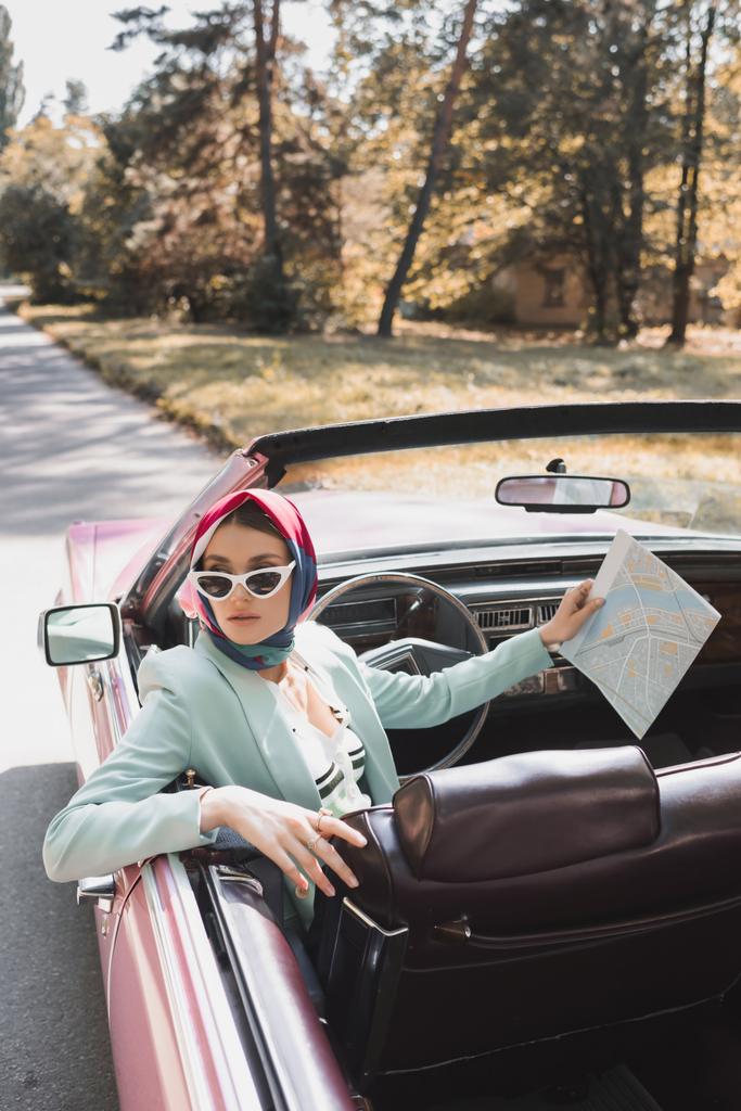 Stylish woman holding map while sitting on driver seat of cabriolet car on road  - Photo, Image