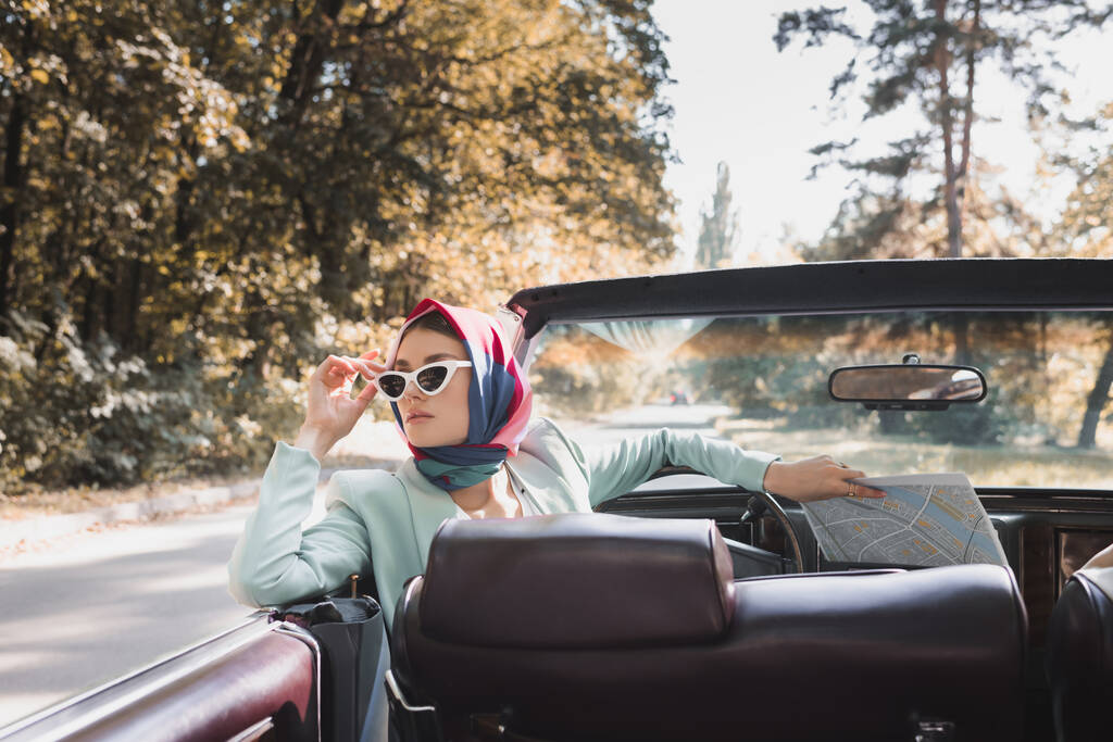 Güneş gözlüklerine dokunan şık bir kadın eski arabanın ön planında haritayı tutuyor. - Fotoğraf, Görsel