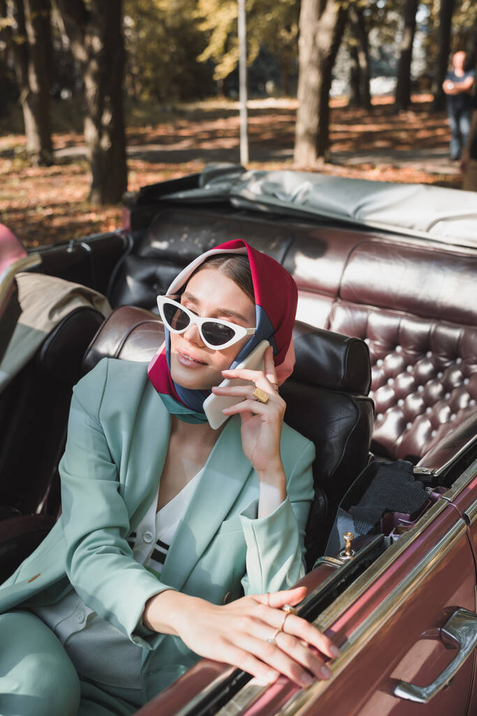Smiling elegant woman talking on smartphone in roofless car during trip  - Photo, Image