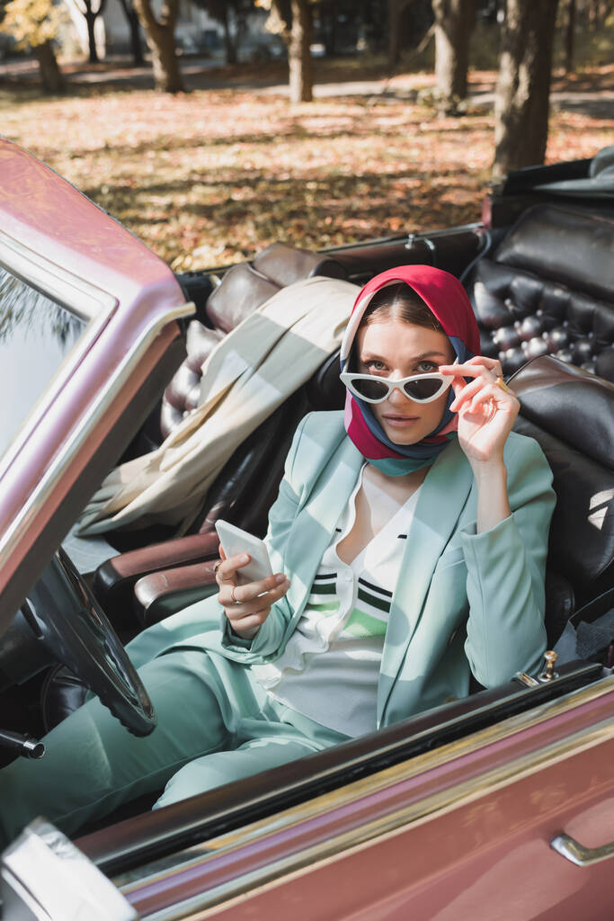 Femme en lunettes de soleil tenant smartphone et regardant la caméra en voiture rétro sur le premier plan flou - Photo, image
