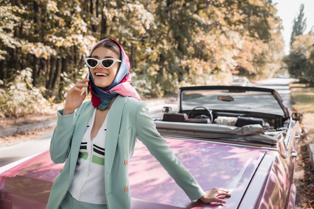Femme souriante en lunettes de soleil parlant sur smartphone près de voiture cabriolet sur la route  - Photo, image