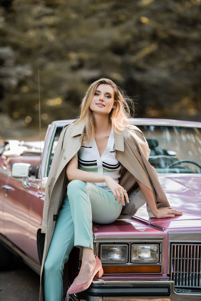 stijlvolle vrouw glimlachen op camera terwijl zitten op de kap van vintage cabriolet - Foto, afbeelding