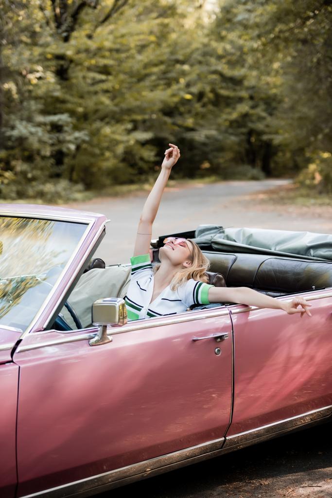 joyful woman relaxing in cabriolet while sitting with raised head, closed eyes and hand in air - Photo, Image