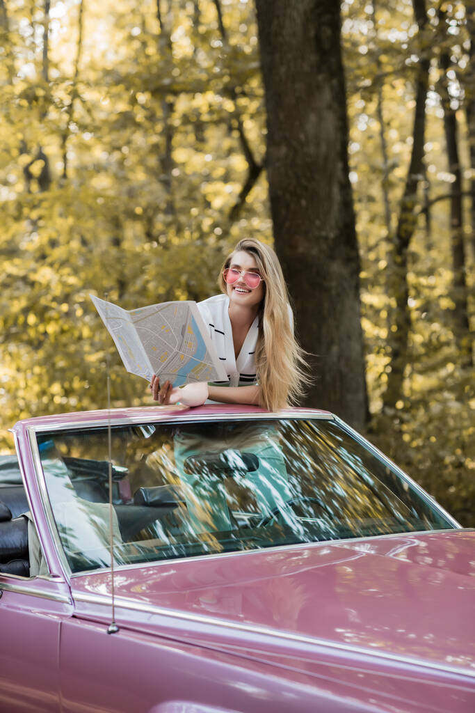 χαμογελαστή γυναίκα με γυαλιά ηλίου κοιτάζοντας οδικό άτλαντα, ενώ κάθεται σε cabriolet στο δάσος - Φωτογραφία, εικόνα